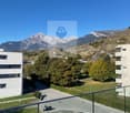 View of parking area with two cars parked, a motorcycle is parked in the shadow of a building, and the building has a balcony with a glass railing overlooking the mountains.
