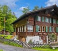 Two-story wooden chalet-style house with green shutters, surrounded by lush greenery and a wooden fence. The house has a covered porch and appears to be in a rural, forested setting.