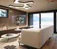 Spacious living room with large windows offering a scenic view of the mountains and lake, modern lighting fixtures, a comfortable beige sofa, a round coffee table, and a wall-mounted TV with various streaming app icons displayed.