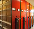 The image shows a row of bright orange storage lockers with shelves above them. The lockers have locks and handles, and there are framed pictures hanging on the wall above the lockers.