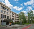 Large multi-story building with a classical architectural style, featuring rows of windows and a covered entrance. The building is surrounded by trees and greenery, with a paved area in front that appears to be a parking lot or plaza. The sky is blue with