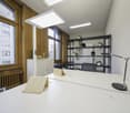 An office space with large windows, white walls, ceiling-mounted lighting, office furniture, radiator, desk lamp, shelves, chairs, and indoor plants.