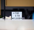 Desk with a lightbox that reads 'THE 5TH FLOOR' in front of a yellow curtain, potted plants on the desk.