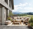 Spacious outdoor patio with wooden deck, comfortable seating area with sofa and armchairs, coffee table, decorative pillows, potted plants, and scenic mountain view in the background