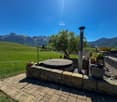 Outdoor patio with a stone fireplace, surrounded by a grassy field and mountains in the background. There is a tree and other landscaping elements visible.