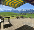 Covered patio with a yellow and green striped awning, overlooking a grassy field and snow-capped mountains in the distance. The patio has a stone tile floor and outdoor furniture, creating a scenic and relaxing outdoor living space.