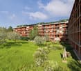 Multi-story apartment building with red and green exterior, surrounded by lush greenery including trees and a grassy lawn, with balconies on each unit