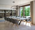 Large open-concept dining area with modern pendant lighting, wooden dining table and chairs, built-in shelving with glassware, and large windows overlooking a garden or outdoor space