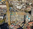Aerial view of building under construction, surrounded by various buildings, cranes, cars parked in lot, and construction equipment scattered throughout site.