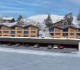 A large building complex with wooden exterior, covered parking area for several cars, snow on the ground