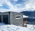 Modern house with stone facade, two garages, wooden balcony, snowy roof, mountainous background