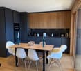 Dining room with wooden table, white chairs, black cabinets, wooden floor, glass door, kitchen area with sink, countertop, and appliances