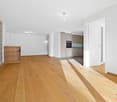 Spacious open-concept living area with hardwood floors, a stone accent wall, and a modern kitchen with built-in appliances including a dishwasher. The room has ample natural light and a clean, minimalist design.