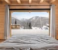 Cozy wooden bedroom with large windows overlooking a snowy mountain landscape. The room features a comfortable bed, wooden beams on the ceiling, and a balcony or terrace with a scenic view of the snow-covered mountains and trees.