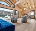 Spacious open-concept living area with high vaulted ceilings, large windows offering a scenic mountain view, wooden walls and floors, comfortable gray sofas and chairs, a dining table, and modern pendant lighting fixtures.