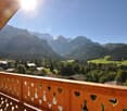 Wooden balcony with heart-shaped cutouts, overlooking a scenic mountain landscape with forests, villages, and snow-capped peaks in the distance under a bright, sunny sky