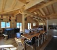 Spacious open-plan living area with high wooden ceilings and beams, large windows providing scenic mountain views, dining table, living room furniture, and a modern kitchen with stainless steel appliances.