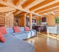 Spacious open-plan living area with wooden beamed ceiling, large windows, hardwood floors, comfortable blue and red sofas, and a kitchen area with dark cabinets and appliances