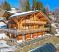 2-story wooden chalet-style house with a large balcony, surrounded by snow-covered trees and hills in a mountainous winter landscape