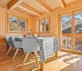 Spacious dining area with wooden walls and ceiling, large windows offering a scenic mountain view, wooden dining table with patterned tablecloth and gray chairs, decorative elements on the table