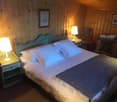 Cozy bedroom with wooden walls, a large bed with blue bedding, two nightstands with lamps, and a framed artwork on the wall. There appears to be a fireplace in the room as well.