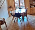 Wooden dining table with chairs, wooden shelves, wooden beams on the ceiling, large window providing natural light, and a fireplace visible in the room.