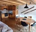 The image shows an open-plan living space with a kitchen, dining area, and living room. The space features wooden beams on the ceiling, a wooden dining table with chairs, a sofa, and a kitchen with wooden cabinets and a dishwasher.