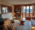 Cozy living room with wooden paneled walls, large windows overlooking a balcony, a fireplace, bookshelves, and a wooden dining table