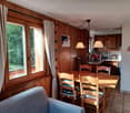The image shows a cozy kitchen and dining area with wooden paneled walls, large windows overlooking a garden, a dining table with chairs, and a kitchen counter with appliances. The room has a warm, rustic feel with decorative elements like wall hangings.