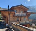 2-story wooden chalet-style house with a balcony, wooden railings, and a chimney. The house is surrounded by mountains and forests, creating a scenic alpine setting.