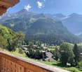 The image shows a picturesque mountain village nestled in a lush, forested valley. The village is surrounded by towering, snow-capped peaks and dense evergreen forests. The wooden balcony or terrace in the foreground offers a panoramic view of the stunnin