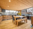 Modern kitchen with wooden flooring, wooden walls, kitchen island with dining table, wooden bench, sliding doors, windows, appliances, television.