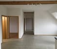 The image shows an empty apartment interior with a hallway leading to multiple rooms. The walls are painted white, and the floors are covered with gray tile. There are wooden beams visible on the ceiling, and the doors leading to the rooms have a wooden f
