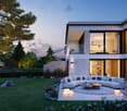 Modern two-story house with glass walls, balcony, outdoor seating area, and garden.