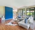 Spacious living room with large windows overlooking a garden, hardwood floors, white leather sofa, blue accent wall, and a modern arc lamp