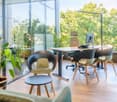 This image shows a modern, open-concept office space with large windows overlooking a lush, green outdoor area. The office features a wooden desk with a computer setup, comfortable office chairs, and a couch. There are also several potted plants throughou