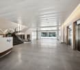 The image shows the interior of a modern office building. The lobby area features a large, open space with a high ceiling, polished tile flooring, and a reception desk with the Regus logo. The space has a clean, minimalist design with neutral colors and p