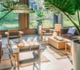 Spacious living room with large windows overlooking a lush, green outdoor area. The room features a mix of modern and natural elements, including wooden furniture, potted plants, and a cozy seating area with sofas and chairs.