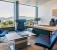 Spacious office with floor-to-ceiling windows offering a panoramic view of the city and mountains. The room features a large wooden desk, comfortable blue swivel chairs, and a coffee table with books. The decor has a modern and minimalist style.