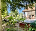 a garden with wooden chairs, tables, and plants, a tree trunk, and a house in the background