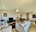 Open-concept living room and dining area, with a large couch, TV stand, and dining table. The kitchen is visible in the background, with modern appliances. The room has large windows, hardwood floors, and a neutral color scheme.