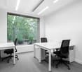 The room has a minimalist design, two desks with black ergonomic chairs, white walls, a yellow accent wall, and a large window with a view of greenery outside.