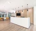 White reception desk, wooden floor, glass walls, large monitor, office chairs, tables, pendant lights.