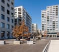 The image shows a modern apartment building complex with several high-rise buildings. The buildings have balconies and large windows. There is a public plaza in the foreground with benches and trees, where people are sitting and relaxing.