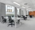 Large open office space with multiple workstations, white tables, black office chairs, and large windows providing natural light. The space has a modern and minimalist design with a gray and orange color scheme.