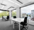 Spacious office with large windows providing a view of the city, multiple workstations with white desks and black office chairs, modern lighting fixtures, and carpeted flooring.