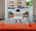 The image shows a modern and stylish interior space with large windows overlooking a street. The space features an orange sofa, two beige armchairs, a round white coffee table, and a black pendant light. The floor is covered with a gray rug, and the walls