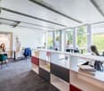 Open-plan office space with large windows providing a view of the surrounding greenery, modern furniture and decor, workstations, and a relaxation area with a chair