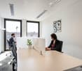 large desk, two people working, long windows, plants, white walls, carpet