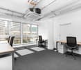 Spacious office space with large windows, providing a bright and open environment. The room features a desk, chairs, and a printer, indicating it is used as a workspace. The carpeted floor and modern furnishings suggest a professional and functional offic